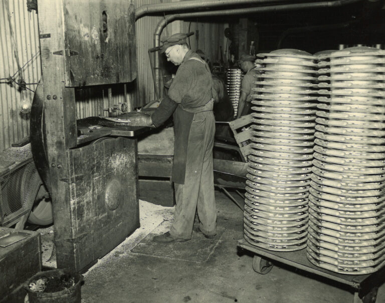 Historic photo of Wisconsin Aluminum foundry worker operating casting equipment, with a stack of automotive parts on a rolling pallet.