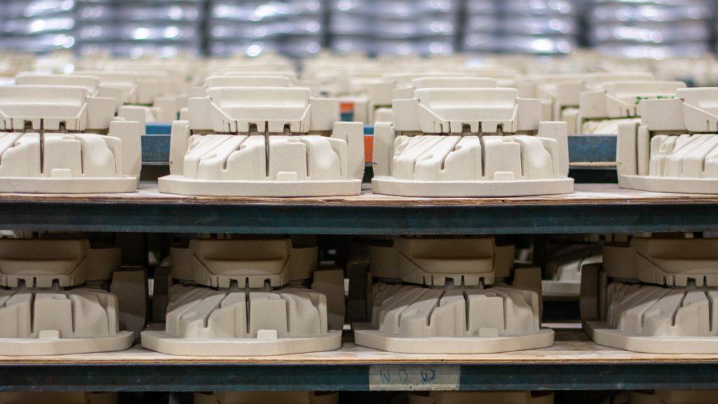 In the foreground, five shelves are stocked with the same complex core, with stacks of cast metal components in the background, slightly blurred.