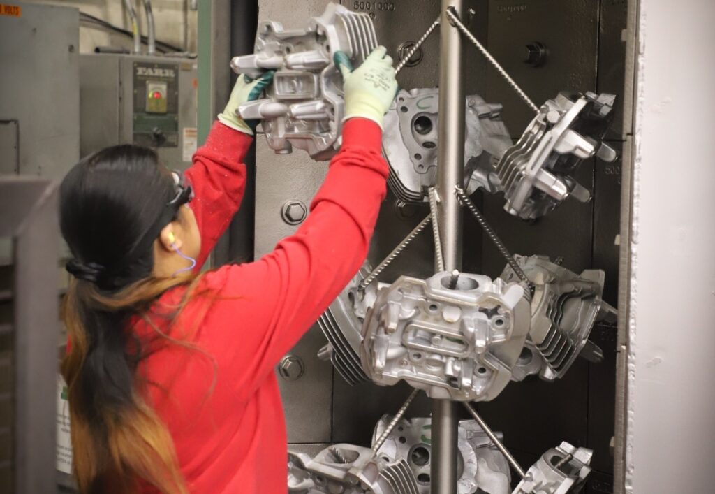 Worker at ATEK Metal Technologies, a Wisconsin Aluminum Foundry company, lifts a cast metal part onto a rod for parts blasting.