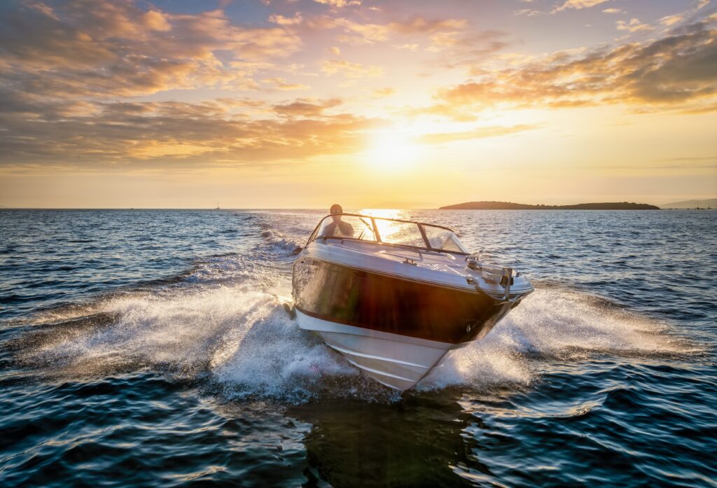 At sunset, a boater drives a speed boat toward camera across open water, creating a wake.